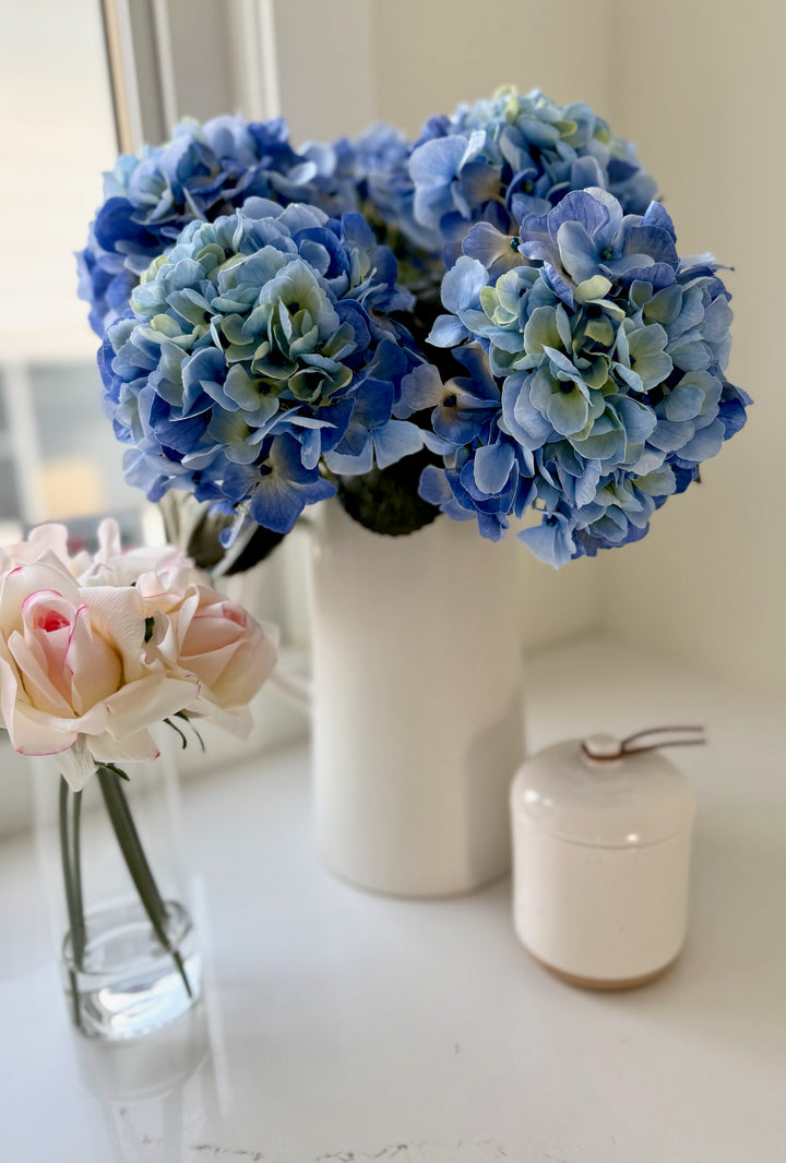 Real touch blue hydrangeas in a white vase on a white surface with a blurred background