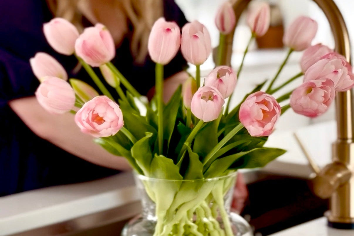 Glass vase with pink tulips on a kitchen counter with a blurred person in the background.