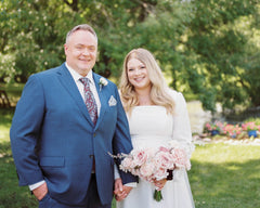 A smiling couple standing outdoors in a garden setting. The man wears a blue suit with a patterned tie and boutonniere, and the woman wears a white wedding dress and holds a pink and white bouquet real touch bouquet. The bouquet is a rental product from the Margot Collection at Eucalypt Co.